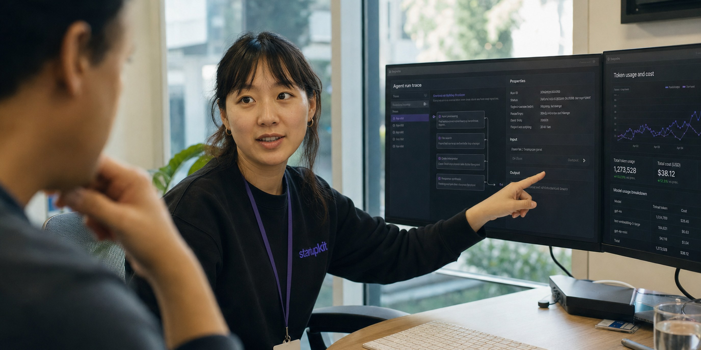 AI engineer being interviewed for a startup role, reviewing an agent run trace and token-cost dashboard on two monitors in a daylit office